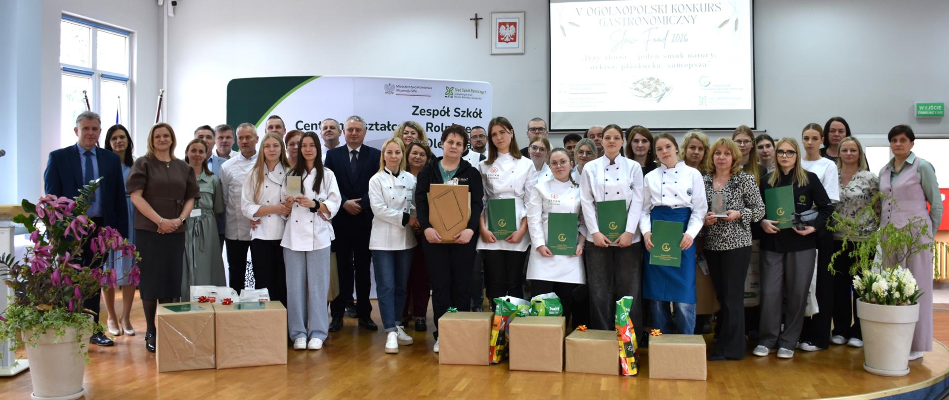 Grupa uczestników, opiekunów i jurorów w strojach kucharskich i formalnych pozuje na scenie podczas gali V Ogólnopolskiego Konkursu Gastronomicznego „SLOW FOOD 2026”.