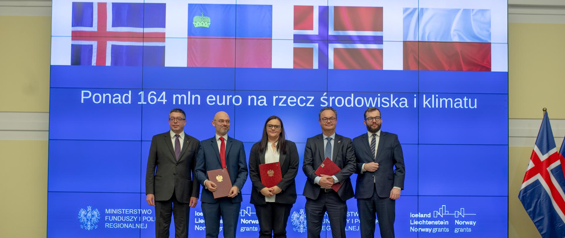 Zdjęcie z podpisanymi umowami, wśród osób znajduje się minister Jarosińska-Jedynak, ambasador Norwegii w Polsce Olav Mykelbust, minister Kurtyka i minister Puda. Za nimi wyświetlany jest napis ponad 164 mln euro na rzecz środowiska i klimatu 
