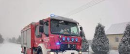 Zdjęcie przedstawiające średni samochód gaśniczy stojący na zaśnieżonej drodze z włączonym oświetleniem zewnętrznym i niebieskimi sygnałami świetlnymi, czerwony kolor karoserii pojazdu, w tle zabudowania i krzewy rosnące przy drodze, widoczny padający śnieg.