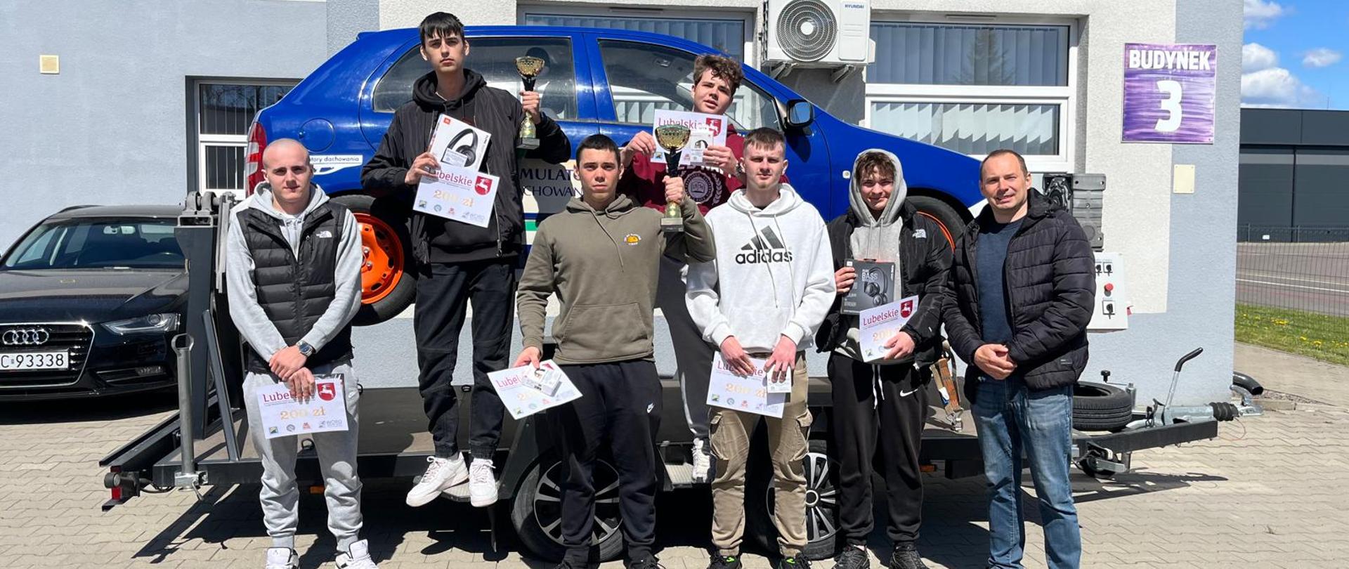 Grupa uczestników z nagrodami i dyplomami stojąca przy samochodzie szkoleniowym przed budynkiem.