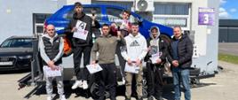 Grupa uczestników z nagrodami i dyplomami stojąca przy samochodzie szkoleniowym przed budynkiem.