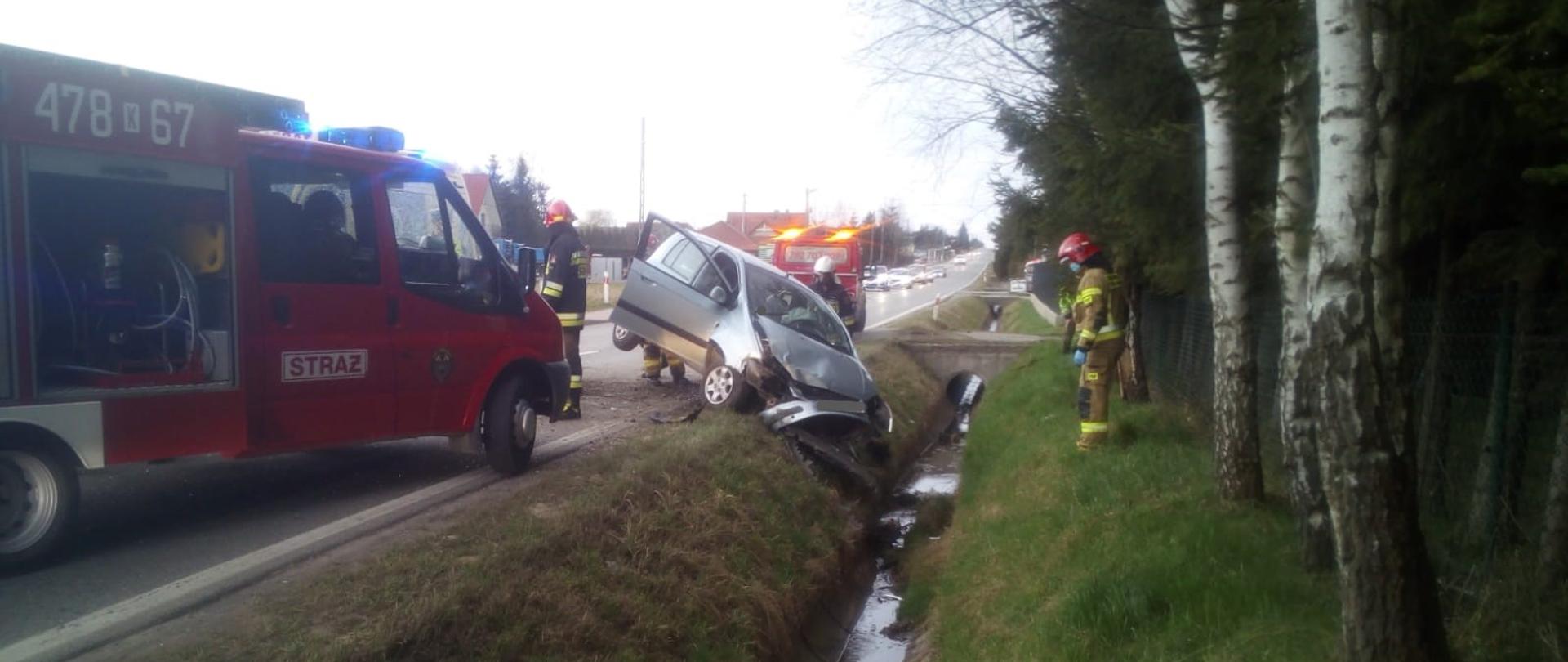 Na zdjęciu w centralnym miejscu widoczny jest samochód osobowy w kolorze srebrnym, który w wyniku uderzenia w betonowy przepust ma zniszczoną przednią część i zawisł nad przydrożnym rowem. Z lewej strony na jezdni znajduje się samochód strażacki. Za rozbitym pojazdem widać samochód pomocy drogowej. Na terenie działań widzimy również strażaków PSP i OSP w umundurowaniu specjalnym i hełmach.