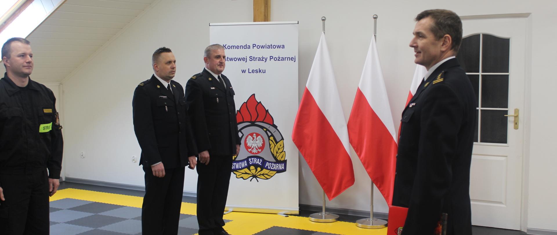 Zdjęcie przedstawia trzech strażaków w mundurach wyjściowych i jednego w mundurze koszarowym Komendy Powiatowej Państwowej Straży Pożarnej w Lesku podczas uroczystej zbiórki z okazji mianowania na stanowisko Dowódcy Jednostki Ratowniczo - Gaśniczej w Lesku oraz na Zastępcy Dowódcy JRG w Lesku. Na zdjęciu Komendant Powiatowy Państwowej Straży Pożarnej w Lesku przemawia do zgromadzonych, stojąc na tle trzech biało- czerwonych flag oraz roll-up-u Komendy Powiatowej Państwowej Straży Pożarnej w Lesku.