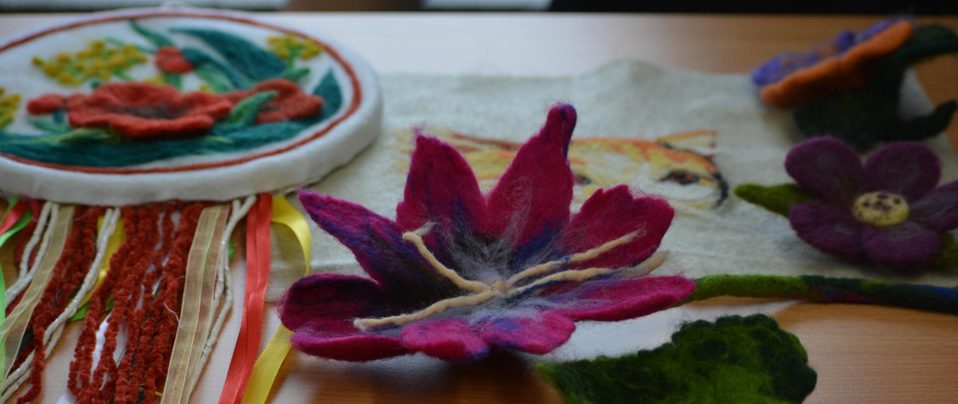 Na drewnianym blacie leżą ozdoby wykonane z wełny. Są tam kwiaty i wyszywanki wykonane techniką filcowania. 
