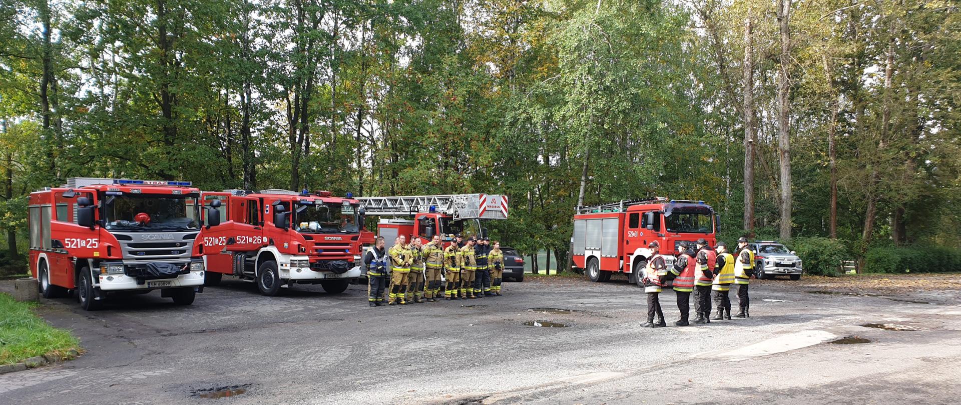 Zdjęcie przedstawia samochody ratowniczo - gaśnicze oraz strażaków stojących na parkingu w otoczeniu drzew, przygotowanych do ćwiczeń. 