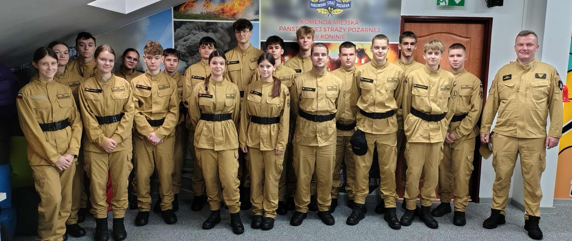 Na zdjęciu widzimy grupę młodych osób ubranych w jednolite, beżowe mundury z czarnymi pasami i butami. Ustawieni są w dwóch rzędach, pozując do zdjęcia grupowego w pomieszczeniu. W tle znajduje się kolorowy baner z napisem „Komenda Miejska Państwowej Straży Pożarnej w Koninie”. Na banerze widoczne są także zdjęcia przedstawiające akcje gaśnicze. 