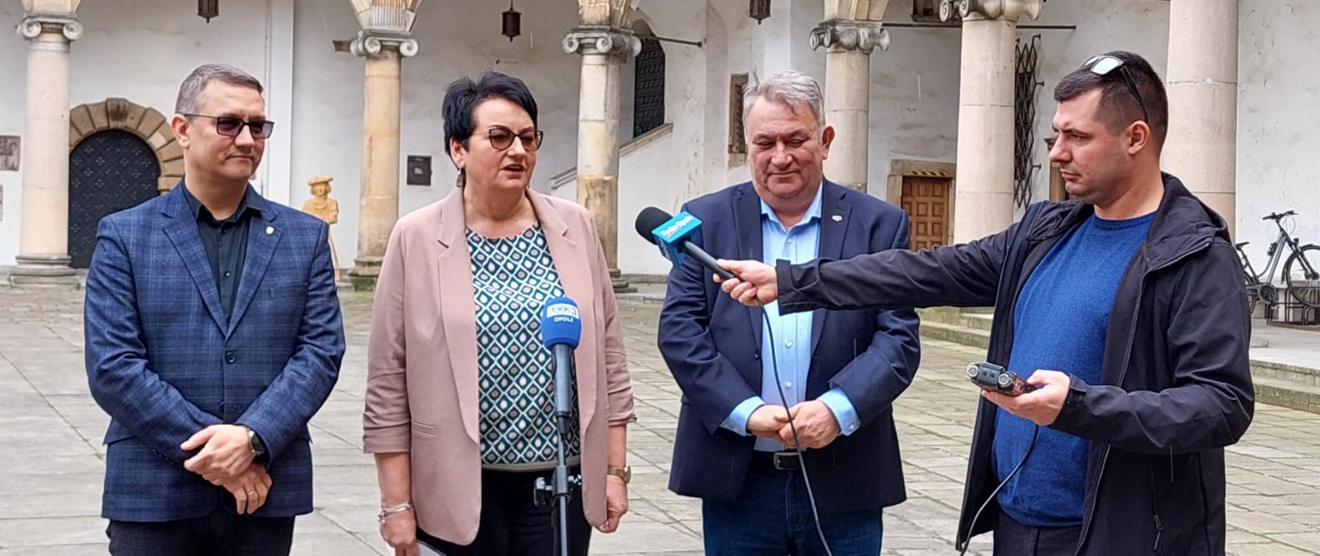 Konferencja prasowa w Muzeum Kresów w Brzegu. Na zdjęciu przy mikrofonach stoi Wojewoda Opolski Monika Jurek, Starosta Powiatu Brzeskiego Jacek Monkiewicz oraz Dyrektor Muzeum Kresów Pan Dariusz Byczkowski. Obok stoi dziennikarz, który trzyma jeden z mikrofonów i nagrywa wypowiedź Pani Wojewody. W tle widać mury muzeum. 