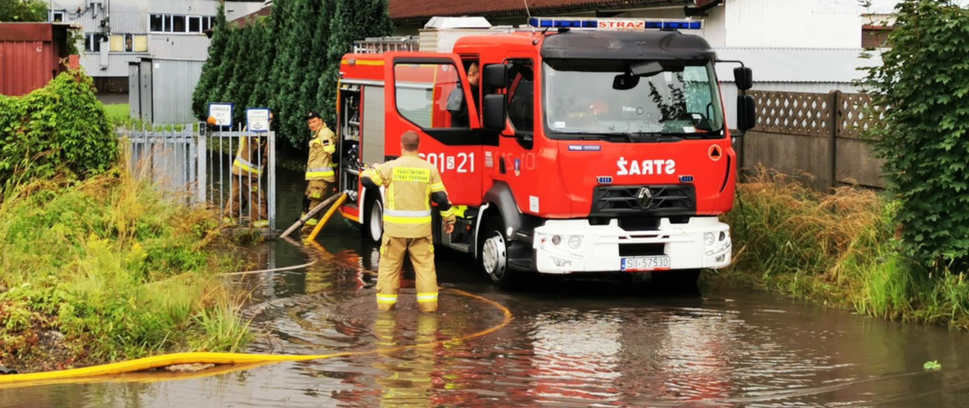 na pierwszym planie rozlewisko wody, na drugim planie stojący w branie samochód ratowniczo-gaśniczy, obok dwóch strażaków ubranych w ubrania specjalne, wypompowujący wodę z rozlewiska