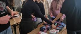 na zdjęciu grupa młodzieży stoi przy stoliku lekcyjnym i bierze ulotki położone na tym stoliku