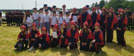 zdjęcie zbiorowe młodzieżowych drużyn Pożarniczych na boisku sportowym z osobami wręczającymi puchary i dyplomy, w tle druhowie OSP