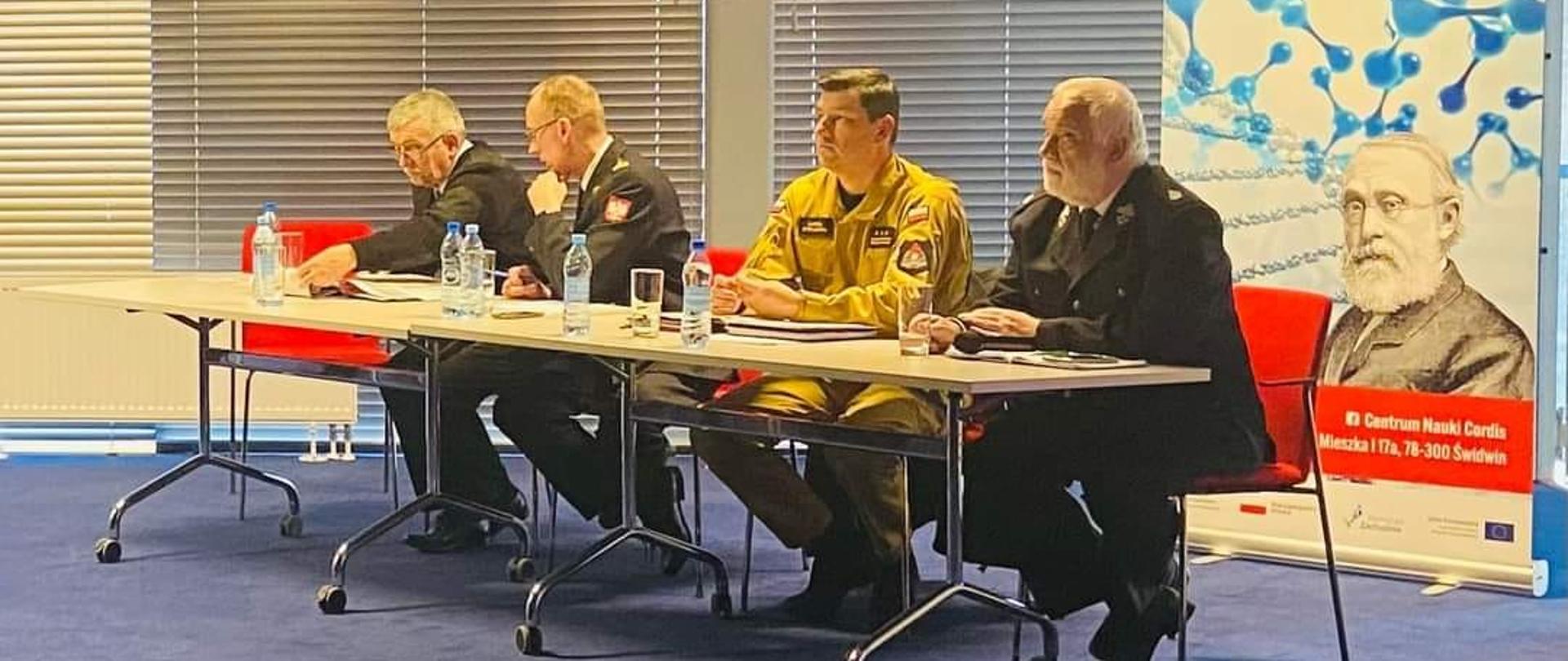 Zdjęcie przedstawia czterech strażaków siedzących przy stole na spotkaniu w sali konferencyjnej.