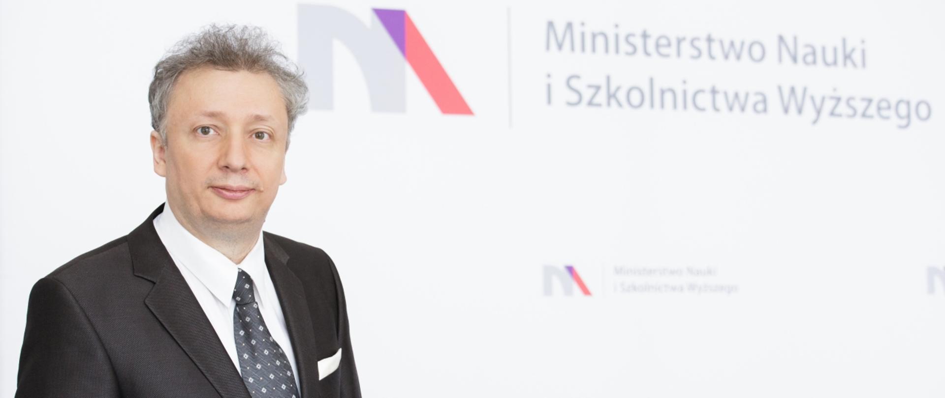 Minister S. Skuza stoi na tle białej ściany z logiem MNiSW