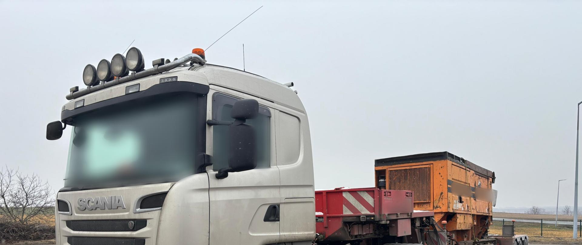 Ciężarówka marki Scania z naczepą niskopodwoziową zaparkowana na placu. Na naczepie przewożona jest duża, pomarańczowa maszyna budowlana; widok z przodu zestawu, pochmurne niebo w tle.
