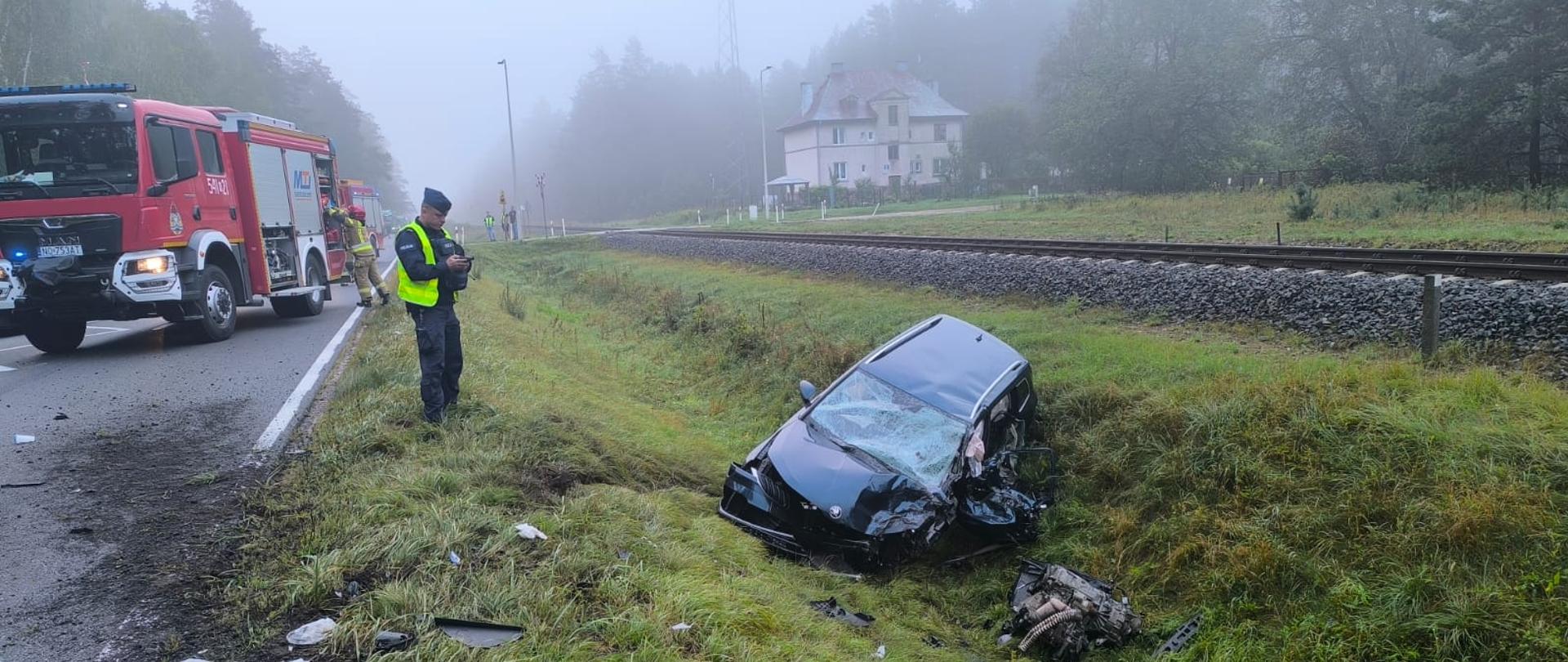 Zniszczony pojazd w rowie, w tle policjant wykonujący czynności, pojazd straży pożarnej oraz przejazd kolejowy, porozrzucane uszkodzone części pojazdu.
