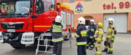Zdjęcie przedstawia strażaków ćwiczących w ramach szkolenia na placu zewnętrznym oraz pojazd strażacki.
W tyle plac komendy i budynek jednostki.
