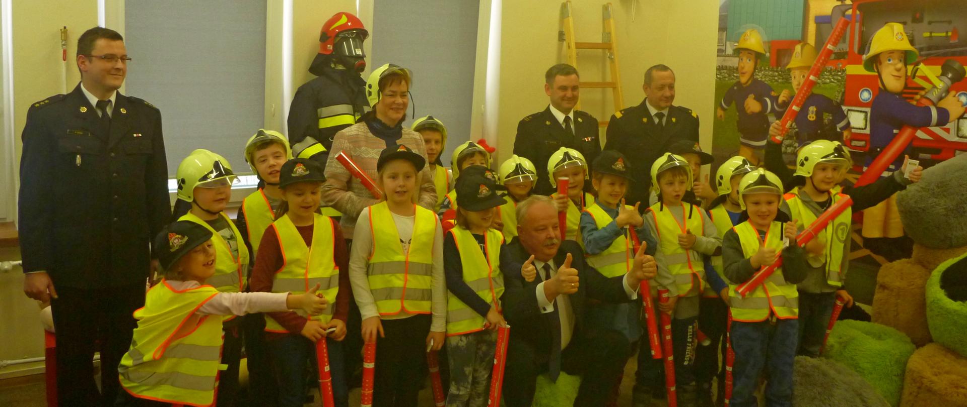 Zdjęcie przedstawia dzieci w kamizelkach odblaskowych na wspólnym podczas otwarcia sali ognik zdjęciu z Panem Posłem Jackiem Osuchem, Małopolskim Komendantem PSP nadbrygadierem Stanisławem Nowakiem, Panią Poseł Lidią Gądek, Z-cą Komendanta Powiatowego PSP w Olkuszu mł. bryg. Michałem Polakiem oraz kpt. Karolem Żabą.