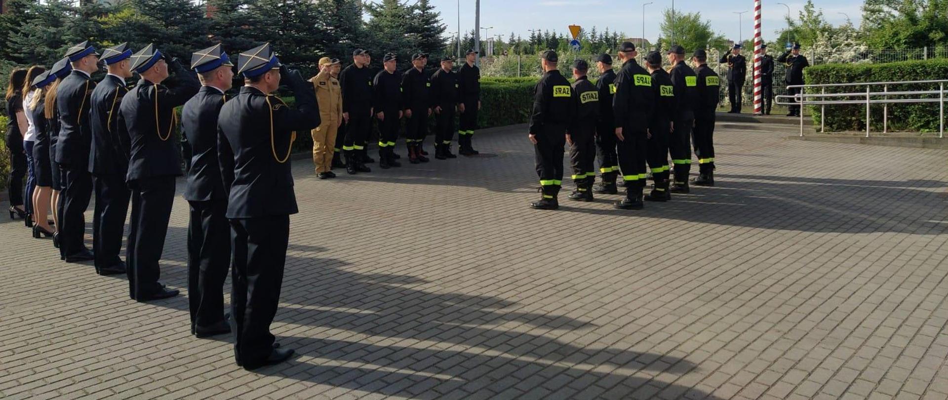 Zdjęcie przedstawia strażaków stojących na placu przed budynkiem komendy podczas uroczystej zbiórki z okazji Święta Flagi