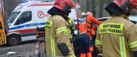Zderzenie auta z koparko-ładowarką. Droga krajowa nr 36 pomiędzy Wodnikami i Załęczem. Na tle ambulansu widoczni są dwaj strażacy i ratownik medyczny, prowadzący działania ratownicze. W tle drzewa.