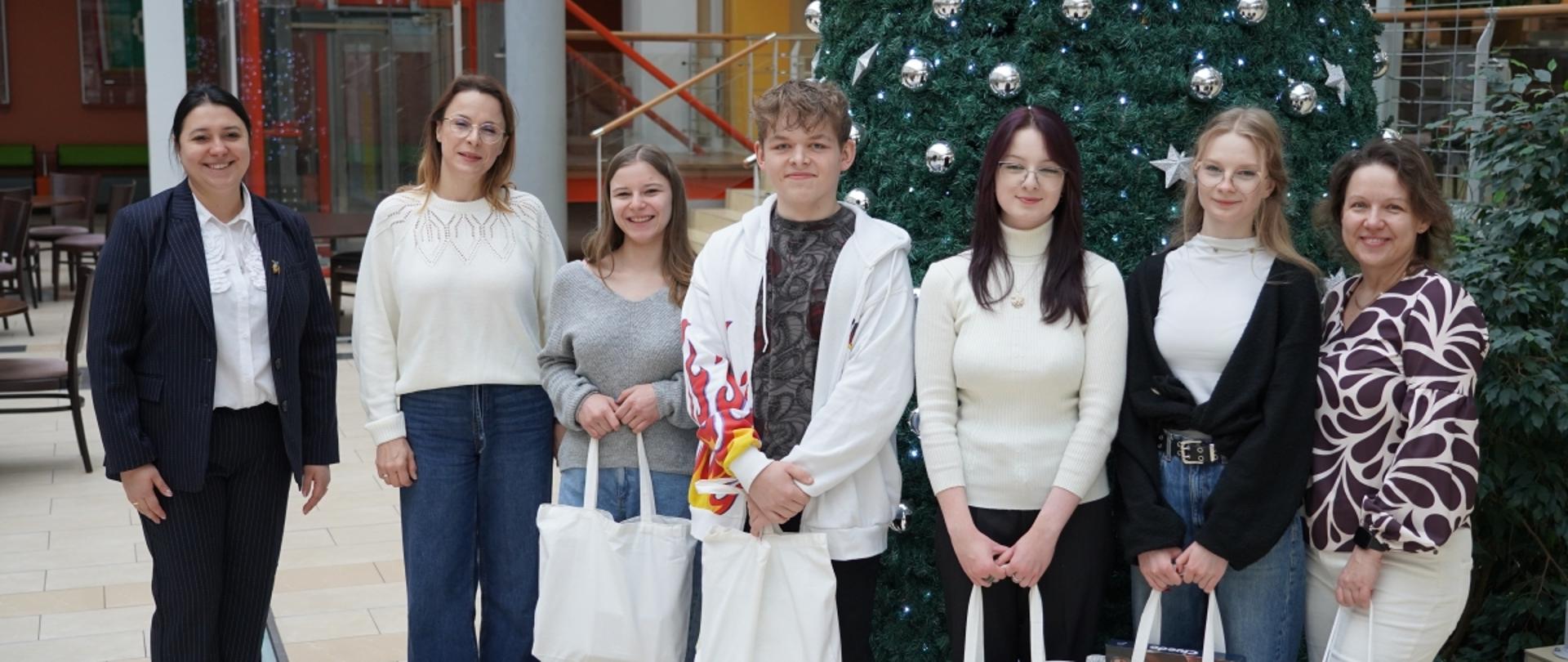 Uczestnicy wydarzenia i organizatorzy pozują do wspólnego zdjęcia przy dużej choince, trzymając białe torby z logo instytucji.