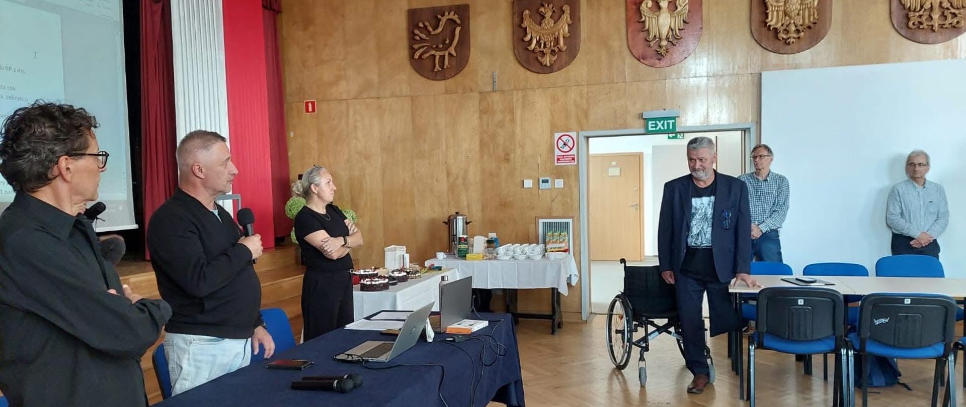 Duże pomieszczenie, kilka osób stoi obok kilku stołów. Jedna trzyma mikrofon, wszyscy patrzą w stronę mężczyzny, który stoi, opierając się ręką o stół