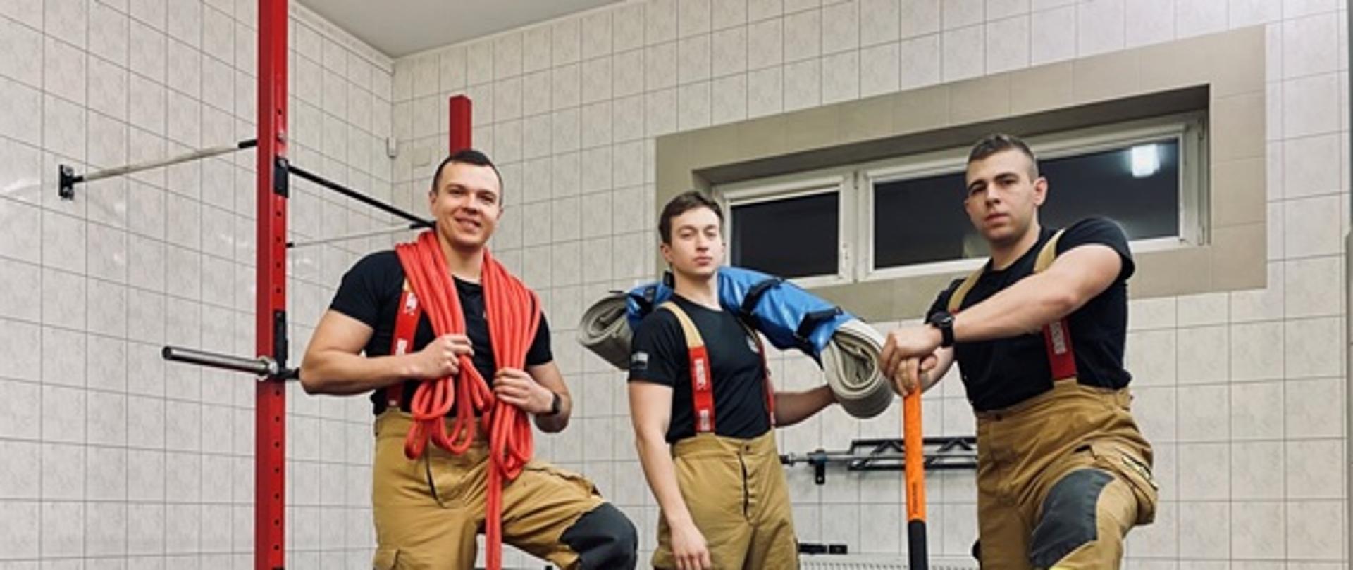 Na zdjęciu widać strażaków. Ubrani w spodnie od ubrania specjalnego w kolorze piaskowym i czarne koszulki. Przed nimi węże pożarnicze w pokrowcach, opona, młot/ Z tyłu widać ściany pomieszczenia z płytkami w kolorze białym. Podłoga ciemna. Po lewej wyposażenie siłowni. Na wysokości głów okno.