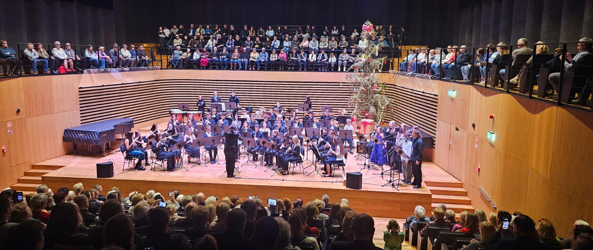 Kobieta dyryguje orkiestrą dętą i zespołem wokalnym. Z przodu i nad sceną widać publiczność.