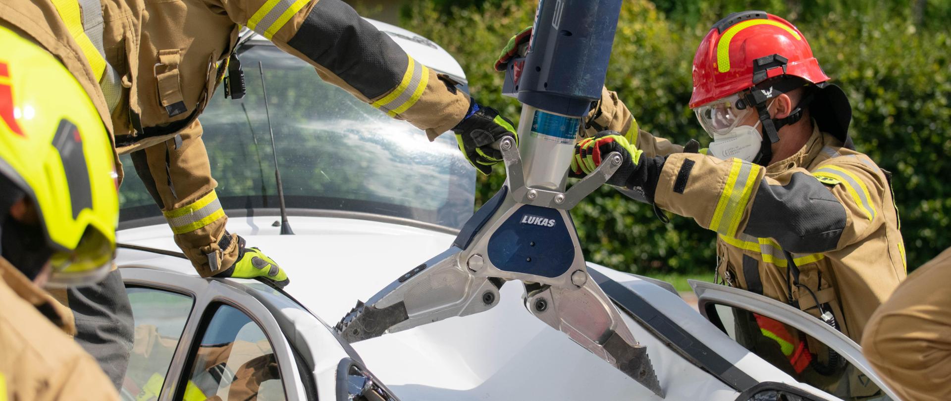 Zdjęcie przedstawia strażaków, którzy rozginają karoserię białego auta po symulowanym wypadku z koparką, przywracając jego elementy do możliwie naturalnych. Samochód jest zniszczony - ma wgięty dach do środka pojazdu, wybitą przednią szybę. W środku znajduje się czerwony manekin.