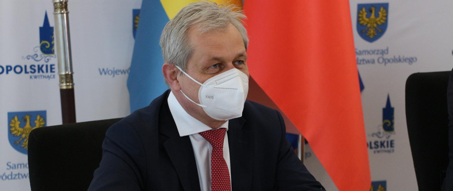 Na zdjęciu siedzący frontalnie ale nieco w ujęciu bocznym wojewoda opolski. Wojewoda siedzi za stołek, w dłoniach trzyma okulary optyczne. Na twarzy ma maseczkę ochronną, spogląda w ustawione przed nim monitory, których nie widać w kadrze. Za jego plecami, nieco po prawej stronie, widać flagę Polski oraz flagę Opolszczyzny oraz ściankę z napisem samorząd województwa. Na dole, na środku kadru widać monitor laptopa.
