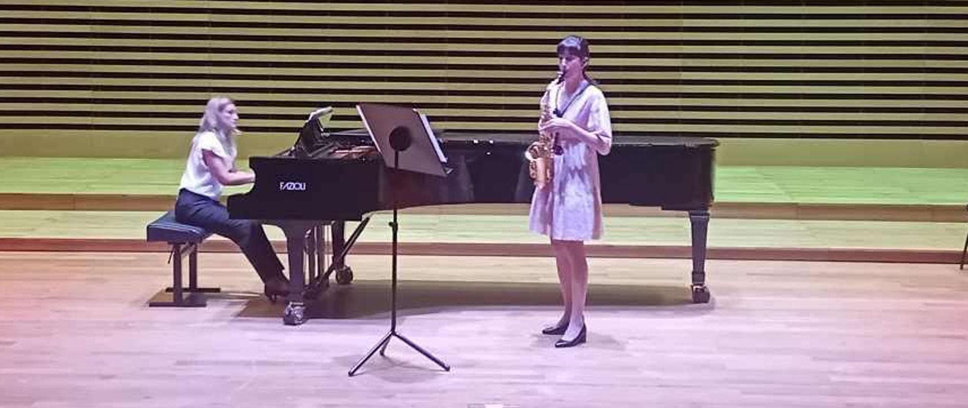 Nastolatka gra na saksofonie, za nią na fortepianie gra kobieta.