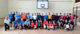 Zdjęcie grupowe osób uczestniczących w turnieju. Wszyscy uśmiechnięci, zadowoleni. Zdjęcie wykonane na hali sportowej. W tle kosz, po prawej drabinki gimnastyczne.