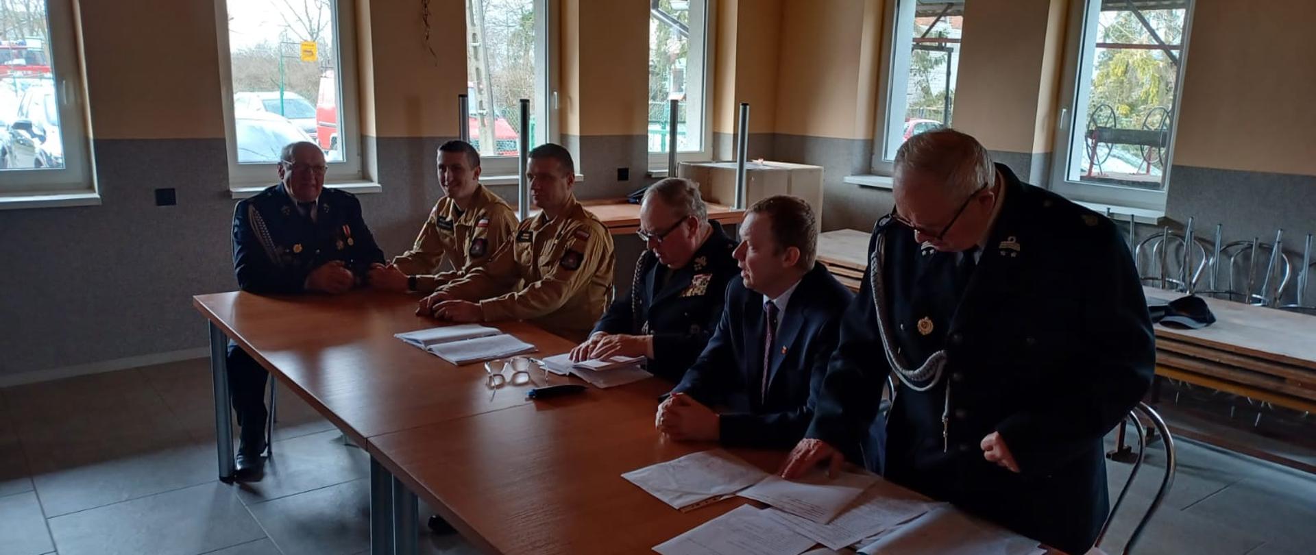 Zdjęcie przedstawia funkcjonariuszy PSP oraz druhów OSP w umundurowaniu wyjściowym na zebraniu Zarządu Oddziału Gminnego ZOSP RP w Osięcinach. Druhowie siedzą na krzesłach w remizie OSP Pilichowo.