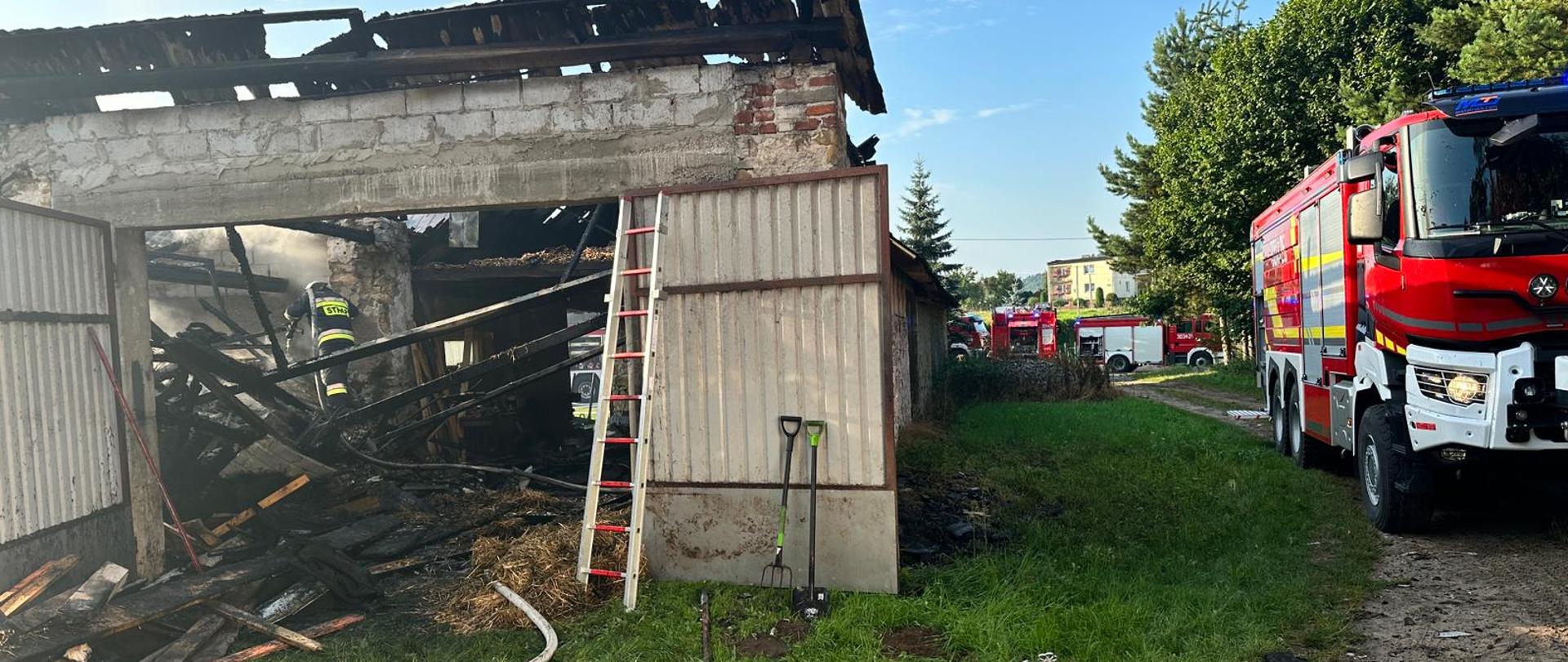 Zdjęcie przedstawia: po lewej stronie budynek gdzie wybuchł pożar. Wewnątrz, przez otwarte wrota widać ratownika podczas kontroli pogorzeliska. Po prawej stronie widać stojące wozy strażackie.