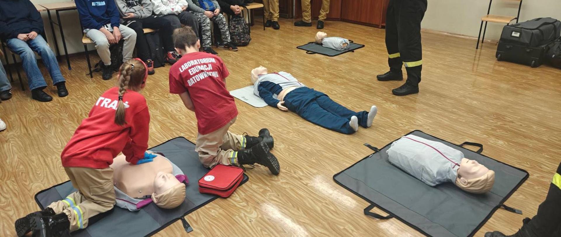 Strażak omawia prowadzenie resuscytacji krążeniowo-oddechowej dla uczniów szkół podstawowych. Dzieci ćwiczą przy fantomach. Pozostali siedzą i obserwują