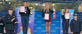 Zdjęcie przedstawia podium zawodów pod niebieskim zadaszeniem. Na wszystkich stopniach stoją młode kobiety. Zawodniczki trzymają w rękach puchary i dyplomy a na szyjach mają zawieszone medale. Po bokach podium stoi 2 strażaków w ciemnych mundurach galowych. W tle niebieskie tło z napisami.