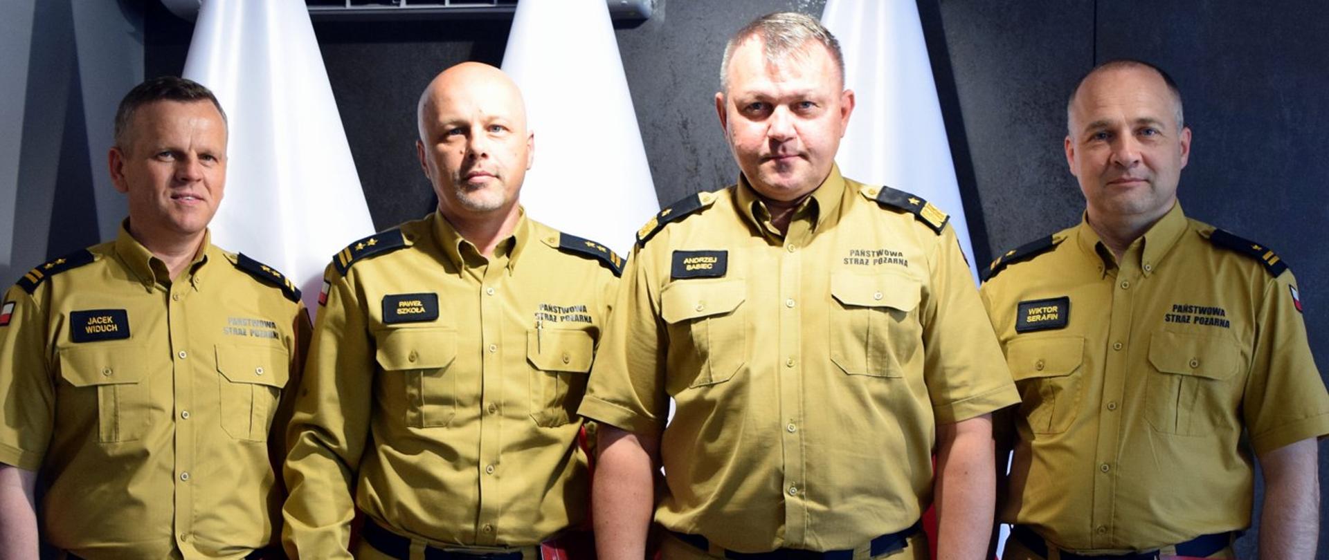 Zdjęcie zrobione wewnątrz pomieszczenia. Na zdjęciu czterech oficerów Państwowej Straży Pożarnej. Za nimi ustawione są biało-czerwone flagi.