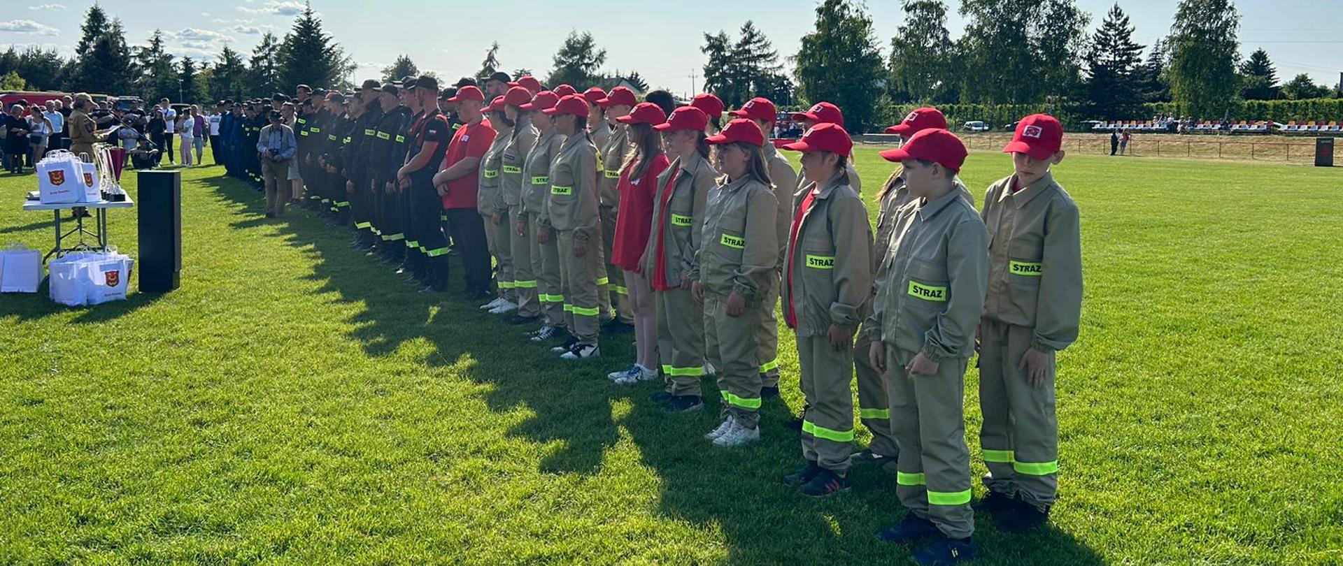 Zdjęcie przedstawia strażaków OSP oraz drużynę MDP ustawionych po prawej stronie w dwuszeregu. Ubrani są w ubranie koszarowe, pasy strażackie oraz hełmy. Strażacy stoją na boisku z trawą.