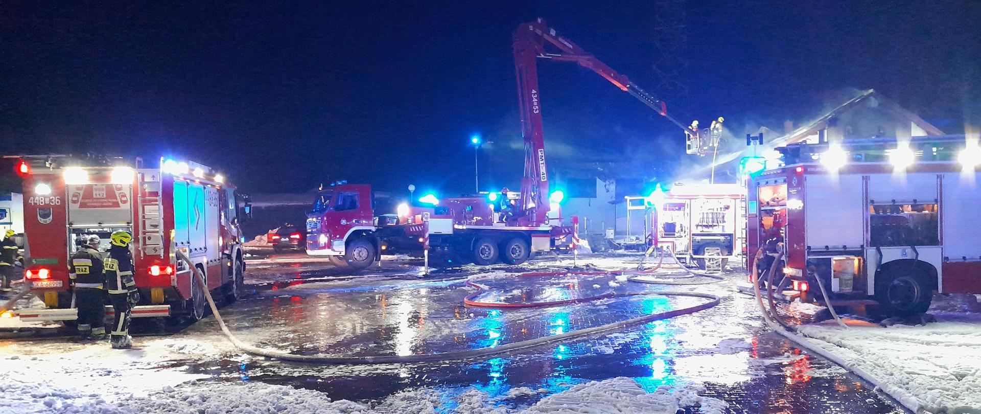 Samochody strażackie na terenie działań gaśniczych ustawione przed budynkiem objętym pożarem.