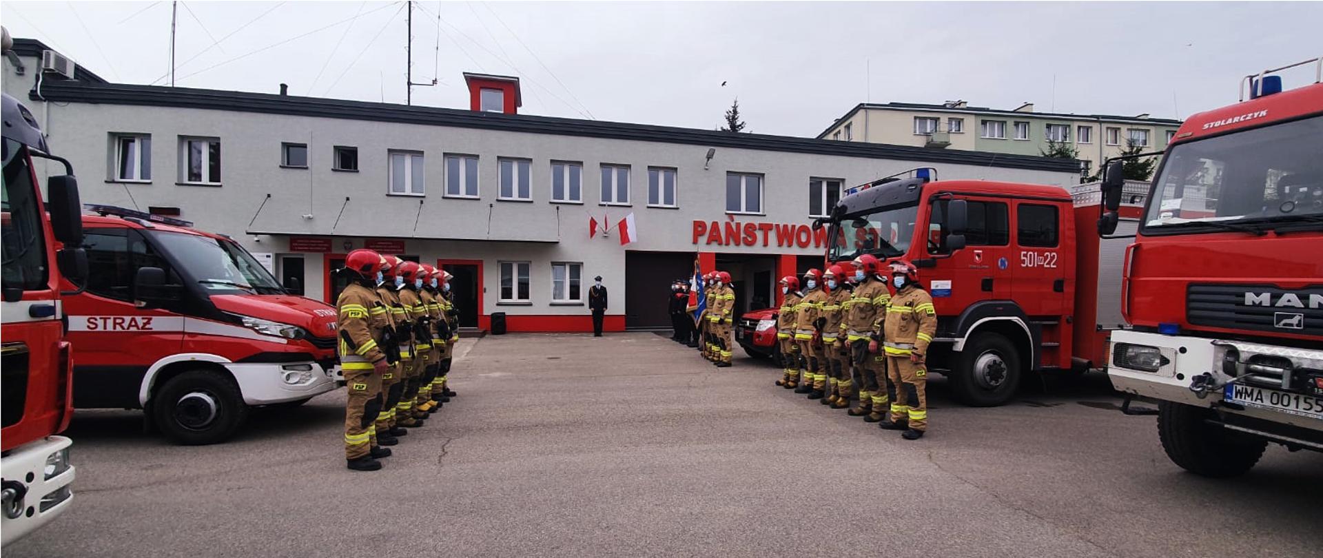Uroczysta zmiana służby przed budynkiem komendy. Dwie zmiany służbowe stoją naprzeciwko siebie przed samochodami strażackimi.