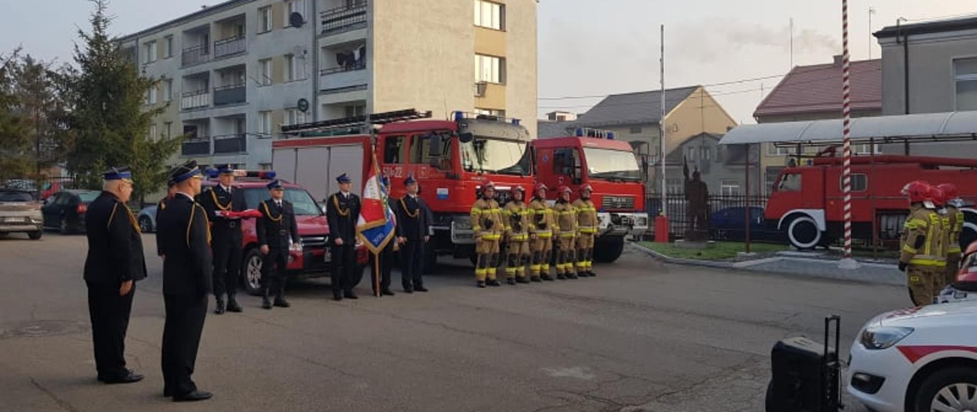 Uroczysta zmiana służby z podniesieniem flagi państwowej na maszt. Poczet flagowy oraz sztandarowy i jedna zmiana służbowa stoją przed samochodami naprzeciw drugiej zmiany służbowej. 