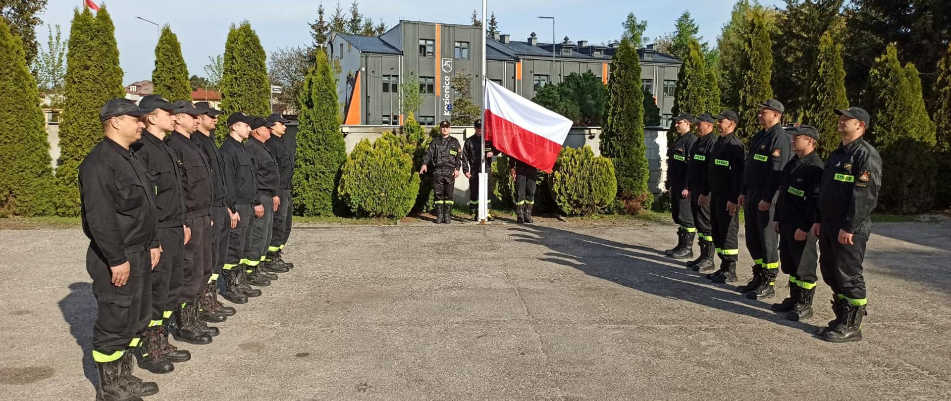 strażacy podczas uroczystej zmiany służbowej w Dzień Flagi Rzeczypospolitej Polskiej stoją na placu komendy , na masz wciągana flaga państwowa