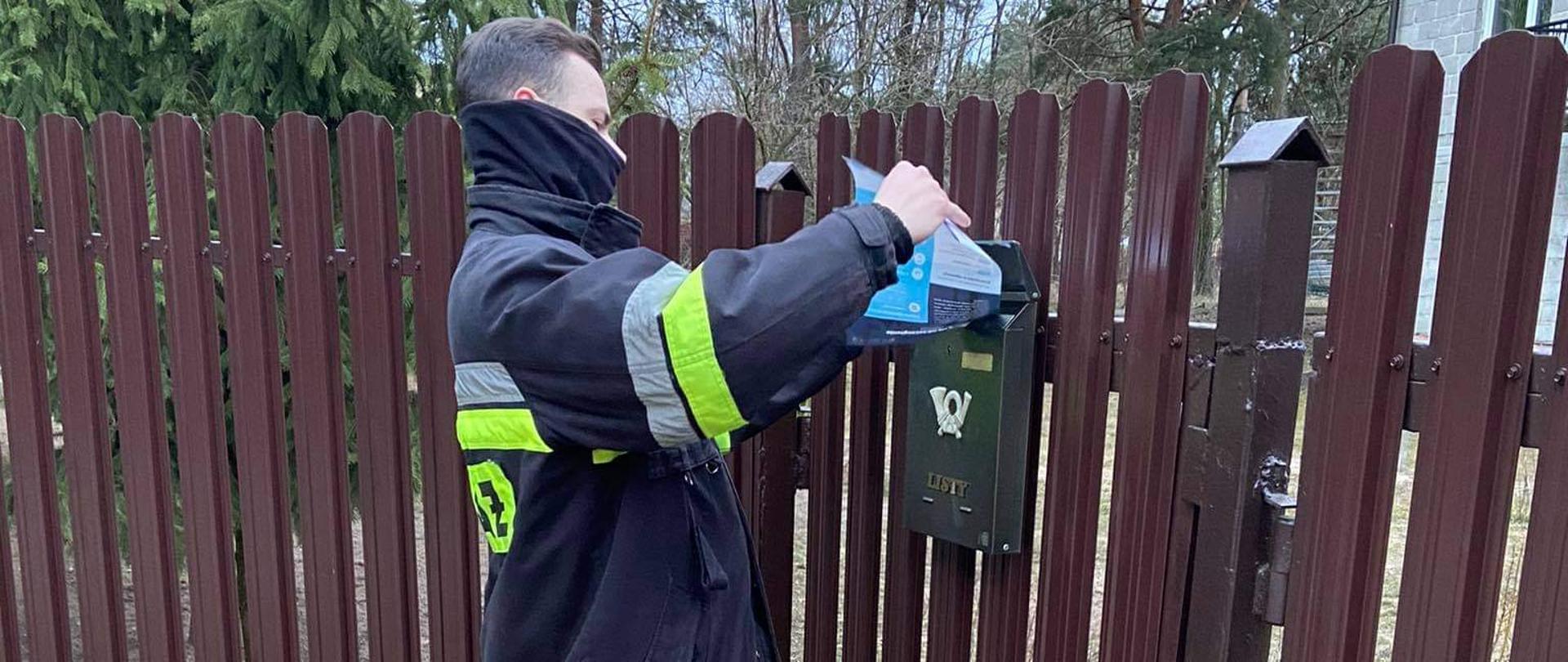 Na zdjęciu druh Ochotniczej Straży Pożarnej ubrany w umundurowanie bojowe wkładający ulotkę do skrzynki na listy zawieszonej na ogrodzeniu posesji.