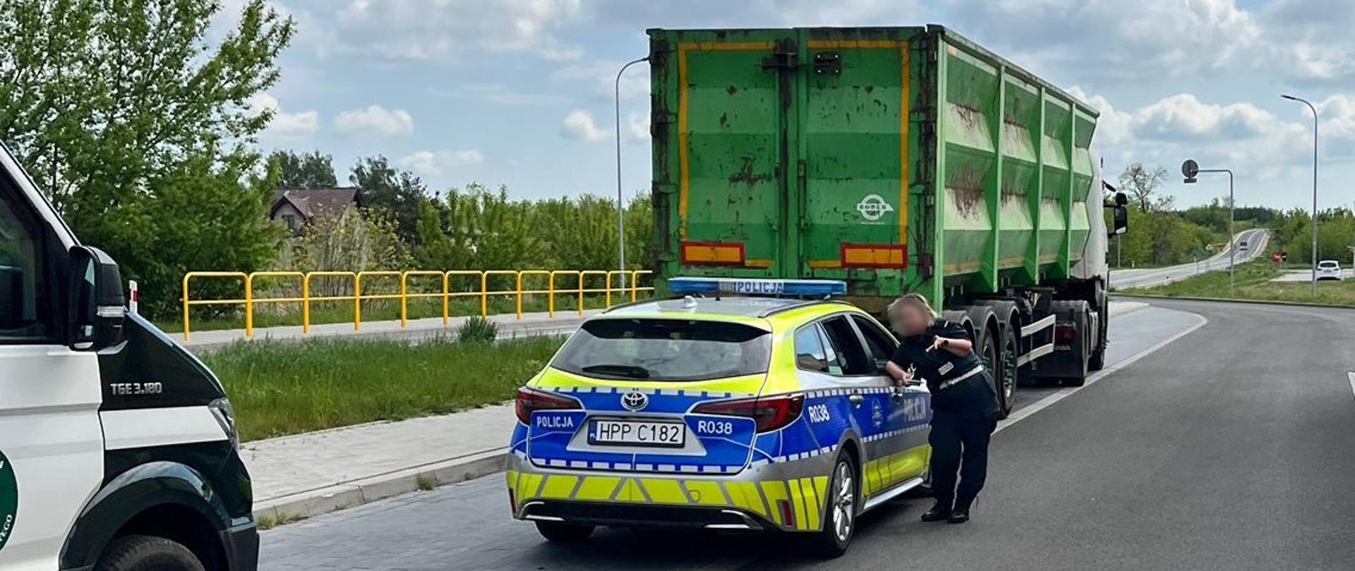 Policyjny radiowóz zatrzymany na poboczu drogi obok ciężarówki z zieloną naczepą do przewozu odpadów. Widoczny jest również fragment przodu radiowozu Inspekcji Transportu Drogowego. Inspektor ITD stoi przy policyjnym radiowozie. W tle widoczna jest jezdnia z żółtymi barierkami, zieleń drzew i krzewów. Widoczne jest błękitne niebo z częściowym zachmurzeniem.