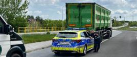 Policyjny radiowóz zatrzymany na poboczu drogi obok ciężarówki z zieloną naczepą do przewozu odpadów. Widoczny jest również fragment przodu radiowozu Inspekcji Transportu Drogowego. Inspektor ITD stoi przy policyjnym radiowozie. W tle widoczna jest jezdnia z żółtymi barierkami, zieleń drzew i krzewów. Widoczne jest błękitne niebo z częściowym zachmurzeniem.