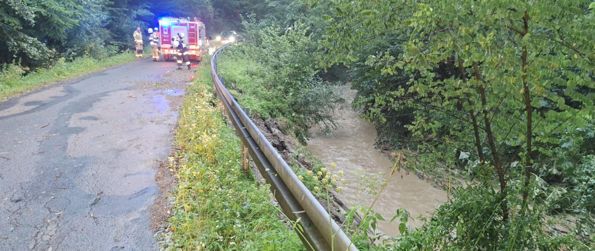 Na zdjęciu widać rwący wezbrany potok po silnych opadach deszczu. W tle samochód pożarniczy.