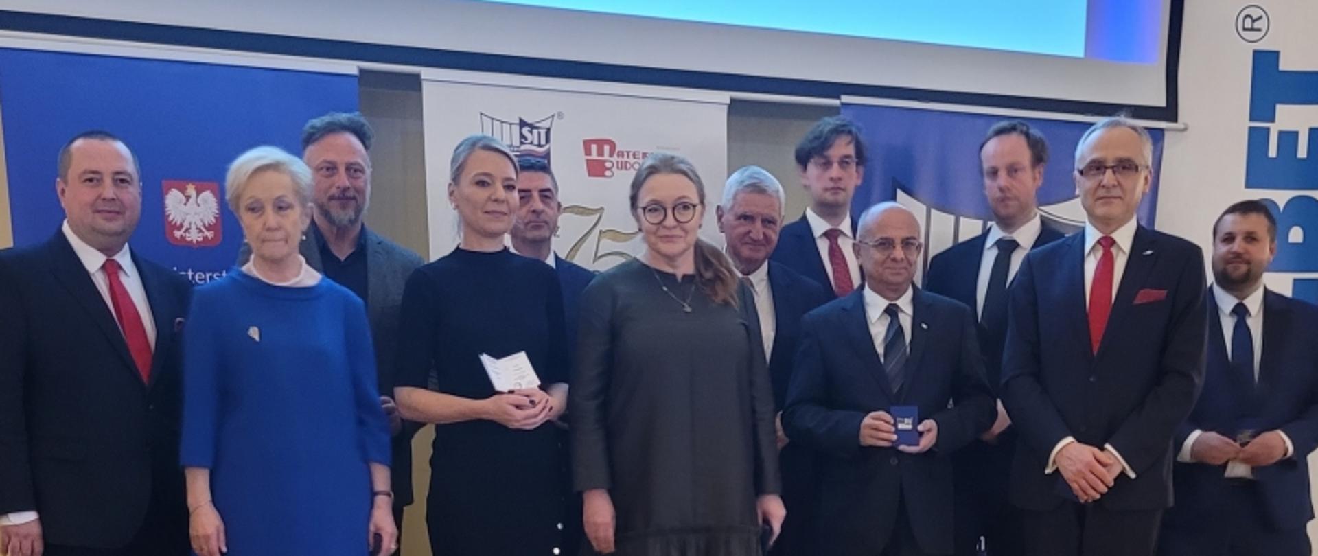 Grupa osób w strojach formalnych stoi przed banerami z logo MZSiT i godłem Polski; jedna osoba trzyma certyfikat.