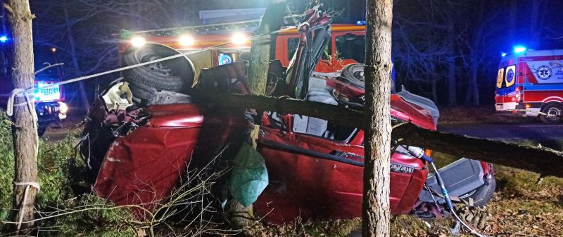 Zdjęcie przedstawia leżące na boku auto. Auto koloru czerwonego. Jest wbite dachem w drzewo. W tle pojazdy służb ratunkowych. Zdjęcie wykonane w nocy.