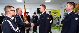 Funkcjonariusz w mundurze przekazał akt włączenia jednostki OSP do KSRG na zdjęciu widać jak poprzez uścisk dłoni dwóch strażaków sobie przekazuje akt. obok stoją dwaj inni strażacy w mundurach w tle sztandar 