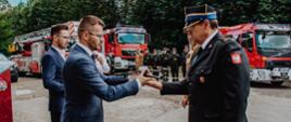 Widok z boku. Na pierwszym planie Burmistrz Wadowic przekazuje drona na ręce Komendanta Powiatowego PSP. W tle samochody pożarnicze oraz załoga na uroczystej zbiórce. 