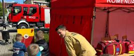 Na zdjęciu widać grupę dzieci oraz strażaka w mundurze, koloru piaskowego. Po prawej stronie znajduje się czerwony namiot, a w jego wnętrzu widoczne są torby ratownicze. W tle zaparkowany jest czerwony wóz strażacki, a za nim stoi budynek z wieżyczką i zegarem, na którego szczycie powiewa polska flaga. Jest widno.