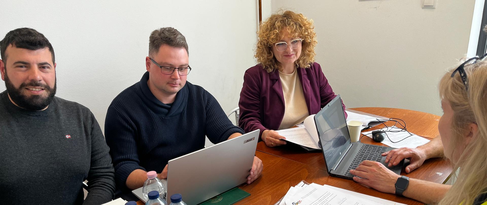 Dwie kobiety i dwóch mężczyzna siedzących przy stole. Na stole dokumenty, komputery oraz butelki z wodą